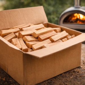 Firewood in a cardboard box with a wood-fired oven in the background.